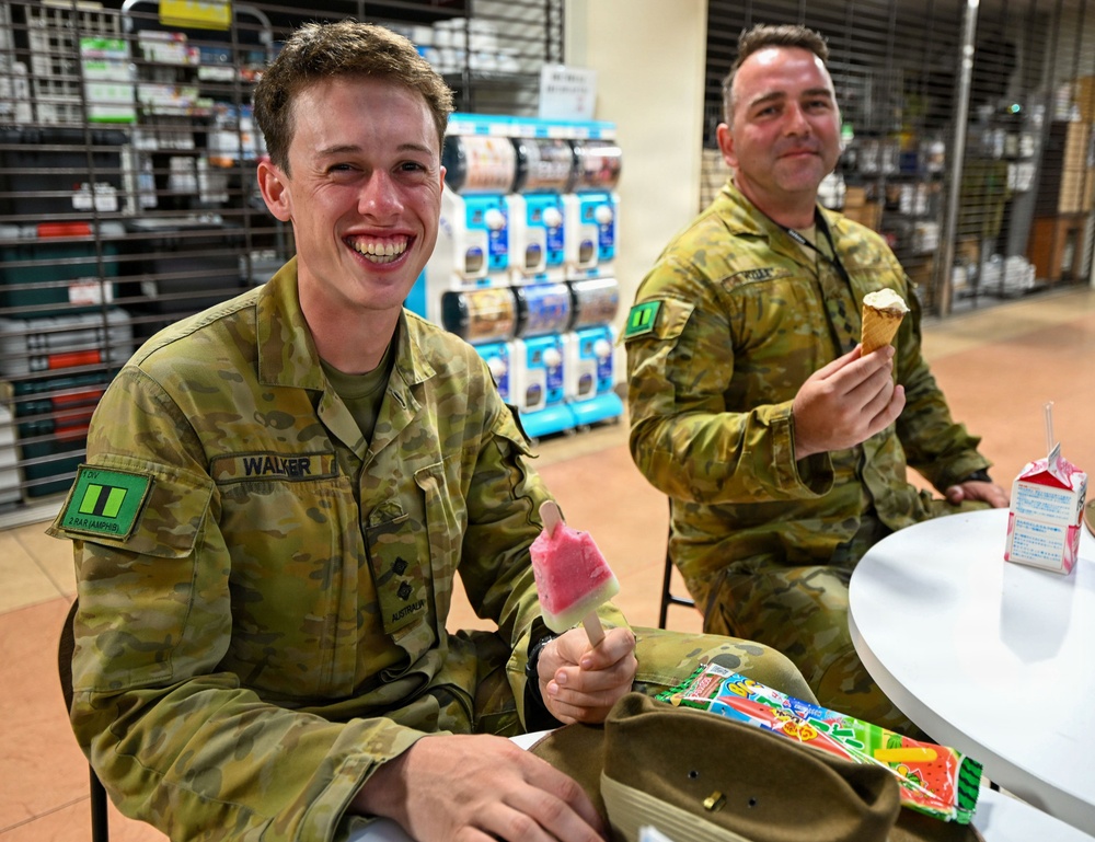Two Australian Army soldiers in MultiCam camouflage uniforms are seated at a white table, enjoying ice cream treats in what appears to be a retail or convenience store setting. The soldier in the foreground, identified by his name tape as 'WALKER,' is smiling broadly while holding a pink/red popsicle, with a bag of Japanese ice cream treats on the table. The second soldier in the background is eating a waffle cone ice cream.