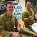 Two Australian Army soldiers in MultiCam camouflage uniforms are seated at a white table, enjoying ice cream treats in what appears to be a retail or convenience store setting. The soldier in the foreground, identified by his name tape as 'WALKER,' is smiling broadly while holding a pink/red popsicle, with a bag of Japanese ice cream treats on the table. The second soldier in the background is eating a waffle cone ice cream.