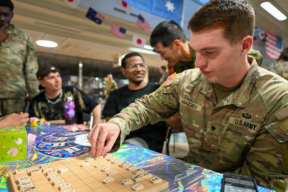 A U.S. Army soldier named Mazzeo is playing Shogi (Japanese chess) at a table surrounded by fellow service members in a multicultural military setting. The room is decorated with international flags including those of the United States, Japan, and Australia, suggesting a multinational military exchange or cultural event. Several other personnel, both in uniform and civilian clothing, are observing or participating in the activity.