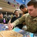 A U.S. Army soldier named Mazzeo is playing Shogi (Japanese chess) at a table surrounded by fellow service members in a multicultural military setting. The room is decorated with international flags including those of the United States, Japan, and Australia, suggesting a multinational military exchange or cultural event. Several other personnel, both in uniform and civilian clothing, are observing or participating in the activity.