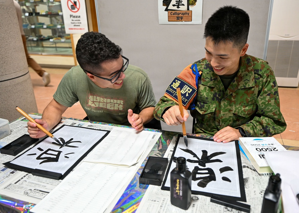 Two young men are engaged in a Japanese calligraphy (習字) activity at what appears to be a cultural exchange event. One person wears a civilian green t-shirt with glasses, while the other is dressed in Japanese military camouflage uniform with an 'Interpreter' armband. Both are smiling and holding calligraphy brushes over large sheets of paper with bold black kanji characters.