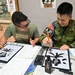 Two young men are engaged in a Japanese calligraphy (習字) activity at what appears to be a cultural exchange event. One person wears a civilian green t-shirt with glasses, while the other is dressed in Japanese military camouflage uniform with an 'Interpreter' armband. Both are smiling and holding calligraphy brushes over large sheets of paper with bold black kanji characters.