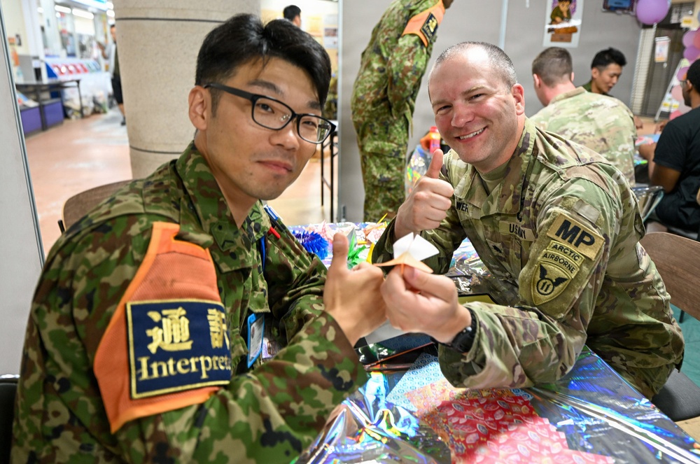 Two military personnel — a Japan Ground Self-Defense Force interpreter and a U.S. Army Military Police soldier assigned to an Arctic Airborne unit — are seated together at a table engaged in origami folding. The U.S. soldier is giving a thumbs-up while both hold small folded paper figures. The setting appears to be an indoor facility, likely during a joint military cultural exchange event.