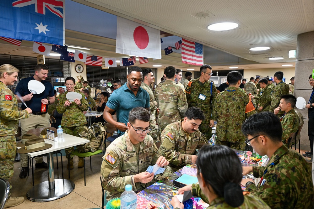 A multinational military gathering is taking place indoors, featuring personnel from the United States, Japan, and Australia, as indicated by the flags displayed overhead. Soldiers in various camouflage uniforms are engaged in what appears to be a cultural exchange activity, with some participants doing origami or paper crafts at a table in the foreground. The event has a friendly, informal atmosphere suggesting a joint military exercise or cultural exchange program.