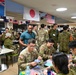 A multinational military gathering is taking place indoors, featuring personnel from the United States, Japan, and Australia, as indicated by the flags displayed overhead. Soldiers in various camouflage uniforms are engaged in what appears to be a cultural exchange activity, with some participants doing origami or paper crafts at a table in the foreground. The event has a friendly, informal atmosphere suggesting a joint military exercise or cultural exchange program.