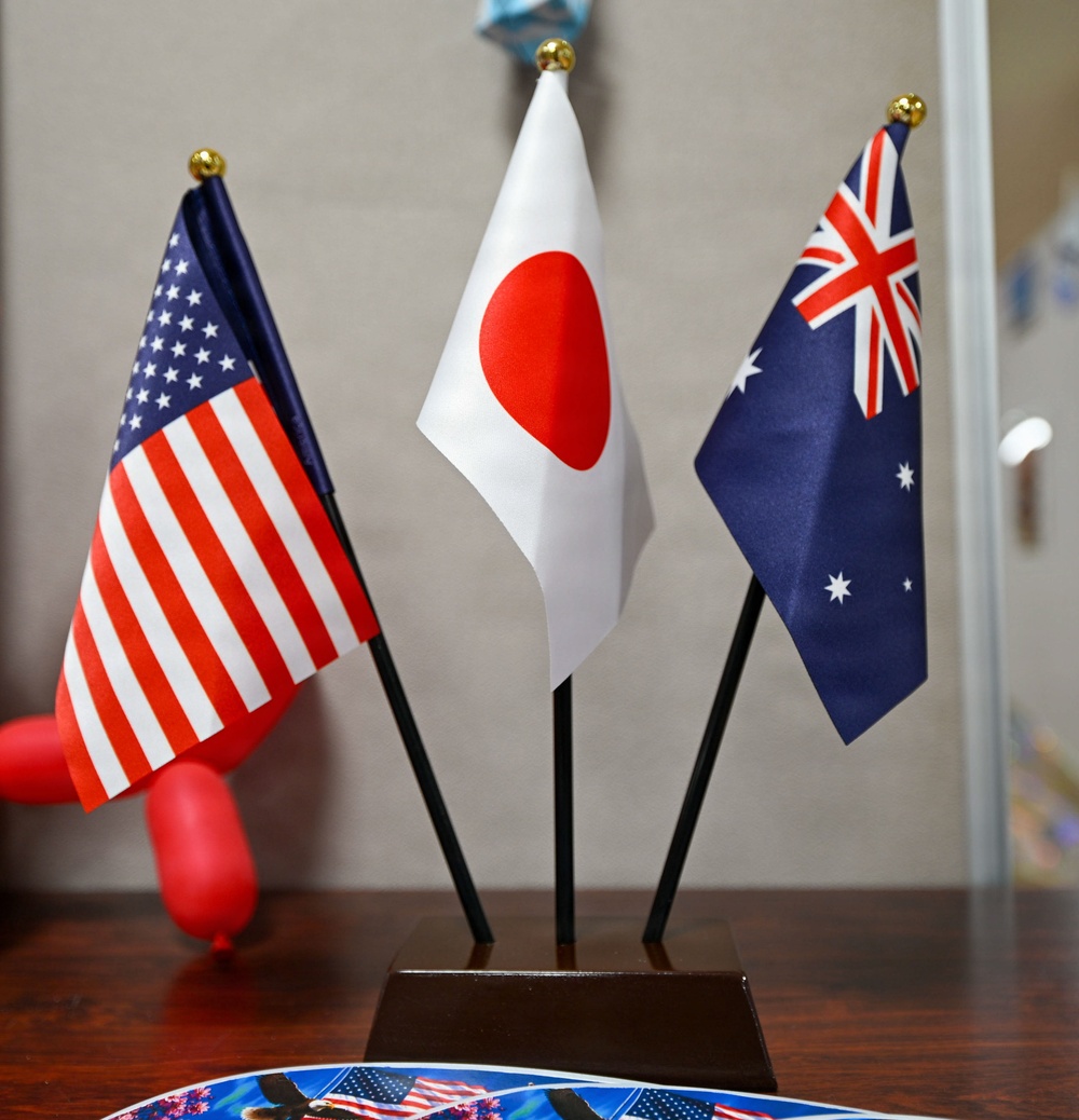 Three small desktop national flags are mounted on a shared dark wooden base with black poles topped by gold finials. The flags represent the United States (left), Japan (center), and Australia (right), suggesting a diplomatic, international, or multilateral context. The display is placed on a wooden surface alongside what appears to be printed materials featuring American imagery.