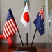 Three small desktop national flags are mounted on a shared dark wooden base with black poles topped by gold finials. The flags represent the United States (left), Japan (center), and Australia (right), suggesting a diplomatic, international, or multilateral context. The display is placed on a wooden surface alongside what appears to be printed materials featuring American imagery.
