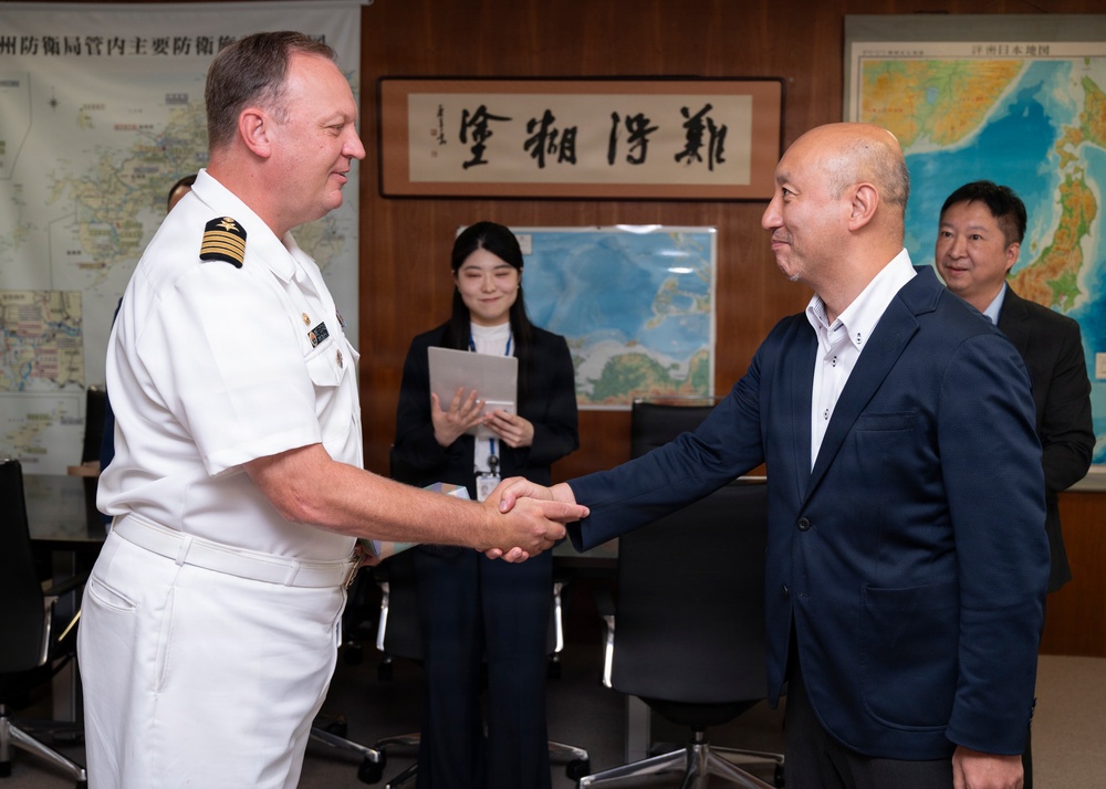 A naval officer in white dress uniform is shaking hands with a civilian man in a navy blazer in what appears to be a Japanese military or government office. The setting includes Japanese maps on the walls and a calligraphy scroll, suggesting an official diplomatic or liaison meeting. Two additional individuals are visible in the background, one holding a tablet or folder.