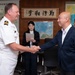A naval officer in white dress uniform is shaking hands with a civilian man in a navy blazer in what appears to be a Japanese military or government office. The setting includes Japanese maps on the walls and a calligraphy scroll, suggesting an official diplomatic or liaison meeting. Two additional individuals are visible in the background, one holding a tablet or folder.
