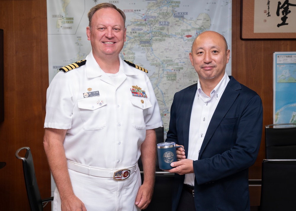 Two men pose together in what appears to be an official office setting. One man is wearing a white U.S. Navy officer's uniform with gold shoulder boards and service ribbons, while the other is dressed in a dark blazer over a white shirt. The civilian is holding a blue commemorative mug, suggesting a formal gift exchange or courtesy visit.