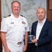 Two men pose together in what appears to be an official office setting. One man is wearing a white U.S. Navy officer's uniform with gold shoulder boards and service ribbons, while the other is dressed in a dark blazer over a white shirt. The civilian is holding a blue commemorative mug, suggesting a formal gift exchange or courtesy visit.