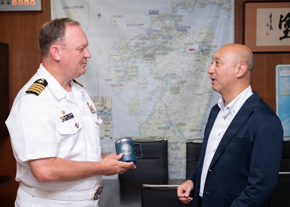 A U.S. Navy officer in white dress uniform is engaged in conversation with a civilian man in a dark blazer, appearing to be in an official meeting or courtesy call setting. The naval officer is holding a dark blue commemorative mug, possibly being presented as a gift. The setting appears to be an official office, likely in Japan based on the Japanese-language map and calligraphy visible in the background.