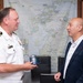 A U.S. Navy officer in white dress uniform is engaged in conversation with a civilian man in a dark blazer, appearing to be in an official meeting or courtesy call setting. The naval officer is holding a dark blue commemorative mug, possibly being presented as a gift. The setting appears to be an official office, likely in Japan based on the Japanese-language map and calligraphy visible in the background.