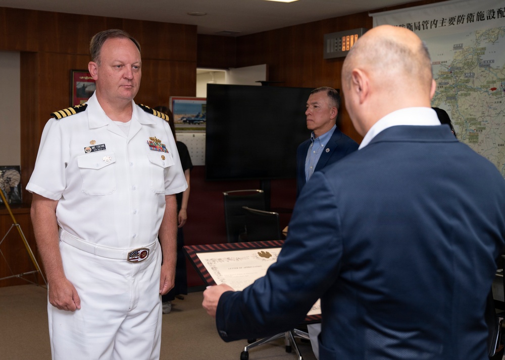 A formal ceremony is taking place in what appears to be a military or government office setting. A U.S. Navy officer in white dress uniform is receiving what appears to be a certificate or letter of appreciation from a civilian in a dark suit. A Japanese map and other office furnishings are visible in the background.