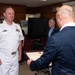 A formal ceremony is taking place in what appears to be a military or government office setting. A U.S. Navy officer in white dress uniform is receiving what appears to be a certificate or letter of appreciation from a civilian in a dark suit. A Japanese map and other office furnishings are visible in the background.