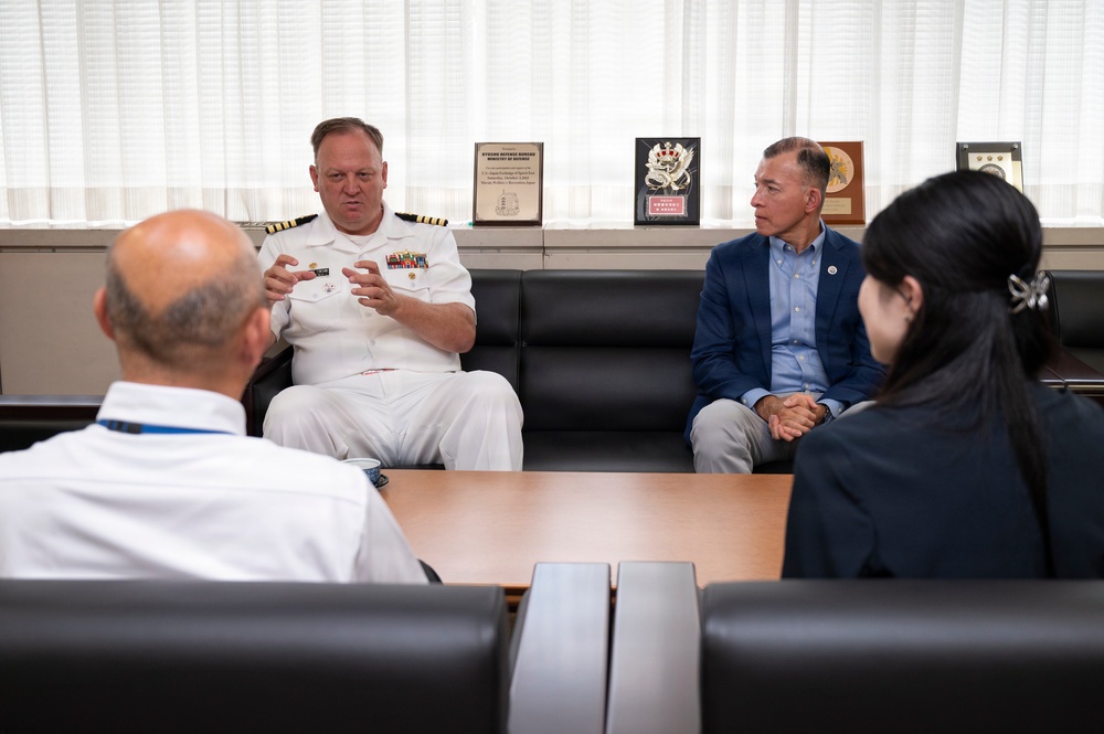 A formal meeting is taking place in what appears to be a military or government office setting. A naval officer in white dress uniform with rank insignia and service ribbons is speaking with several civilians and another uniformed individual. Plaques and awards visible in the background suggest an official diplomatic or institutional environment.