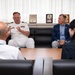 A formal meeting is taking place in what appears to be a military or government office setting. A naval officer in white dress uniform with rank insignia and service ribbons is speaking with several civilians and another uniformed individual. Plaques and awards visible in the background suggest an official diplomatic or institutional environment.