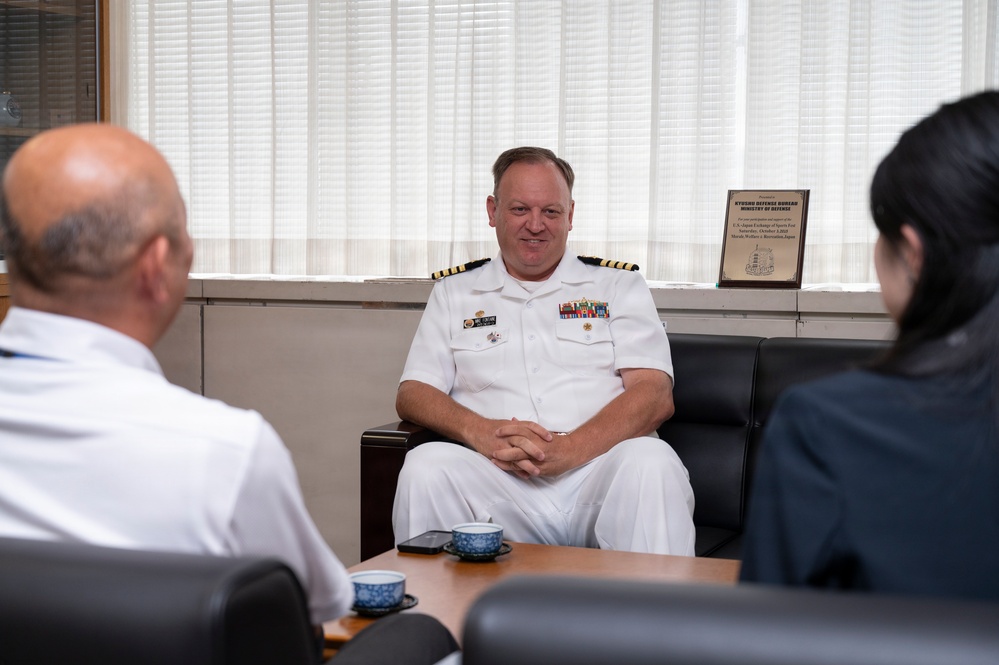 A U.S. Navy officer in white dress uniform is seated and engaged in a meeting or discussion with two other individuals whose backs are visible in the foreground. The setting appears to be an official office environment, with a plaque visible in the background reading 'Kyushu Defense Bureau, Ministry of Defense.' Traditional Japanese tea cups are visible on the table, suggesting a formal diplomatic or liaison meeting.
