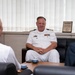 A U.S. Navy officer in white dress uniform is seated and engaged in a meeting or discussion with two other individuals whose backs are visible in the foreground. The setting appears to be an official office environment, with a plaque visible in the background reading 'Kyushu Defense Bureau, Ministry of Defense.' Traditional Japanese tea cups are visible on the table, suggesting a formal diplomatic or liaison meeting.