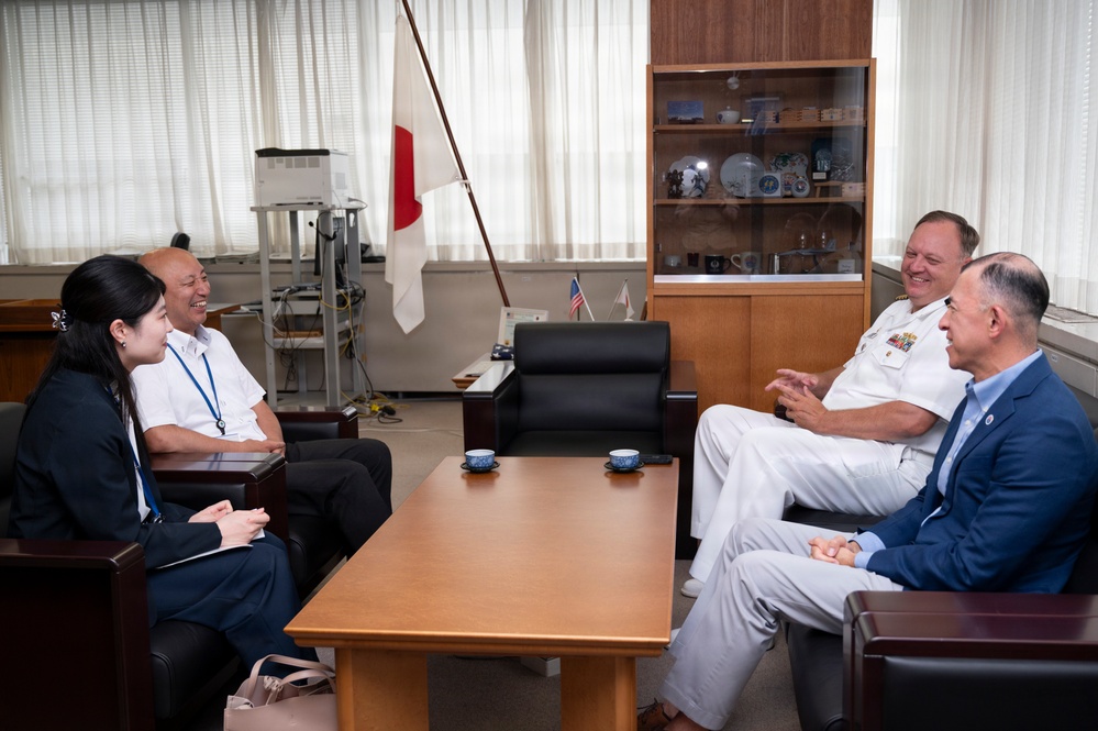 Four individuals are seated in a formal office setting engaged in a meeting or discussion. A U.S. Navy officer in white dress uniform is present alongside three civilians, with Japanese and American flags visible in the background. The atmosphere appears cordial and diplomatic in nature.