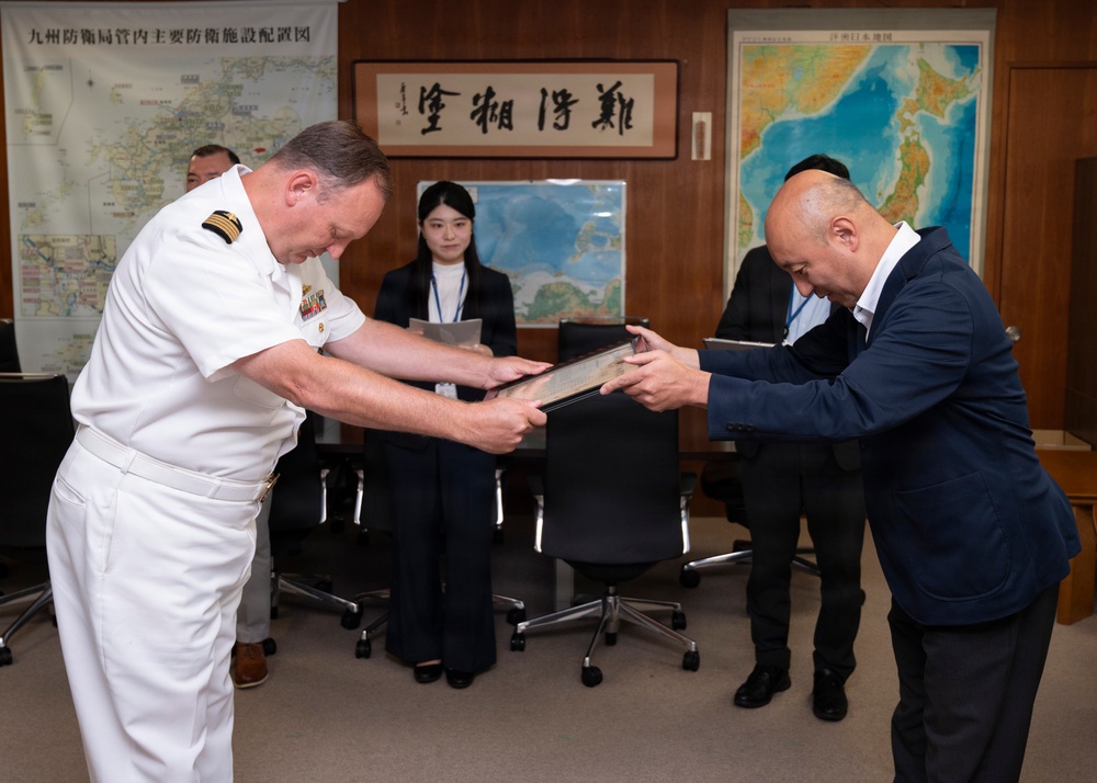 A formal exchange ceremony is taking place in what appears to be a Japanese military or government office, where a naval officer in white dress uniform is exchanging a framed item or document with a civilian official. Both individuals are bowing slightly during the exchange, consistent with Japanese ceremonial protocol. The setting includes maps of Japan and Kyushu on the walls, along with a calligraphy scroll.