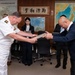 A formal exchange ceremony is taking place in what appears to be a Japanese military or government office, where a naval officer in white dress uniform is exchanging a framed item or document with a civilian official. Both individuals are bowing slightly during the exchange, consistent with Japanese ceremonial protocol. The setting includes maps of Japan and Kyushu on the walls, along with a calligraphy scroll.
