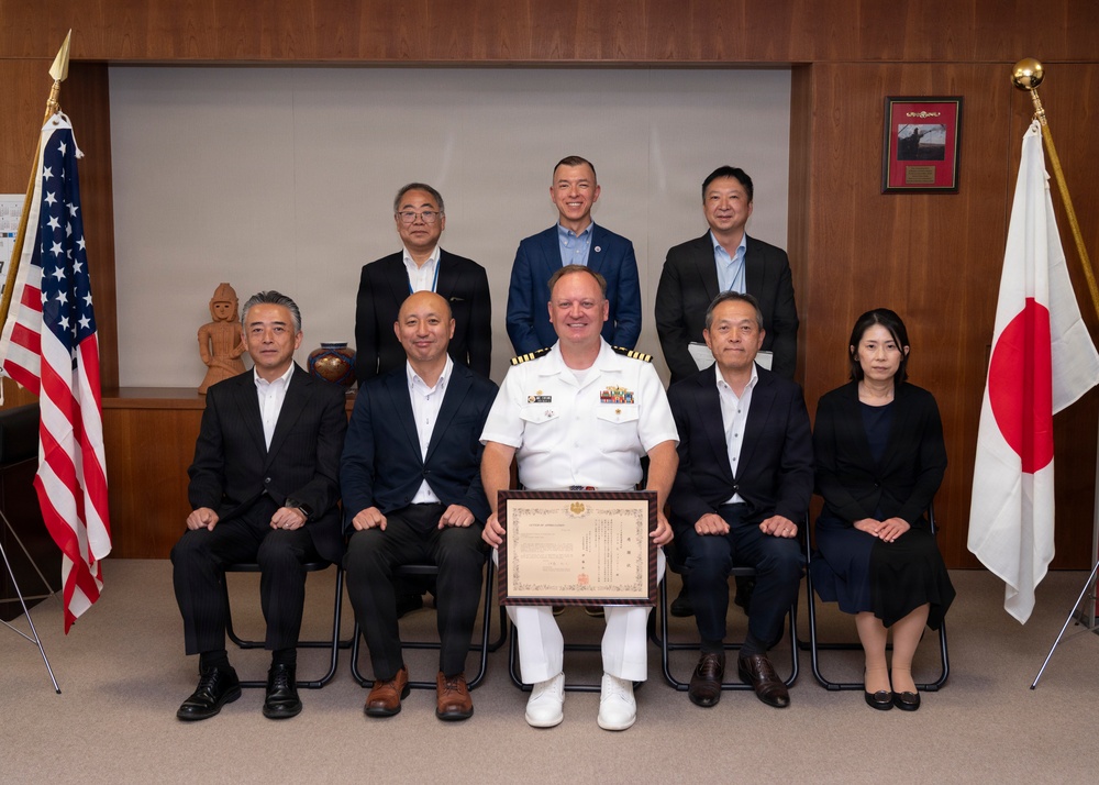 A formal group photograph featuring a U.S. Navy officer in white dress uniform holding a framed certificate or commendation, flanked by eight Japanese civilians in business attire. The setting appears to be an official meeting room with both the American and Japanese flags prominently displayed on either side of the group. This appears to be a bilateral ceremony or award presentation symbolizing U.S.-Japan cooperation.