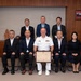 A formal group photograph featuring a U.S. Navy officer in white dress uniform holding a framed certificate or commendation, flanked by eight Japanese civilians in business attire. The setting appears to be an official meeting room with both the American and Japanese flags prominently displayed on either side of the group. This appears to be a bilateral ceremony or award presentation symbolizing U.S.-Japan cooperation.