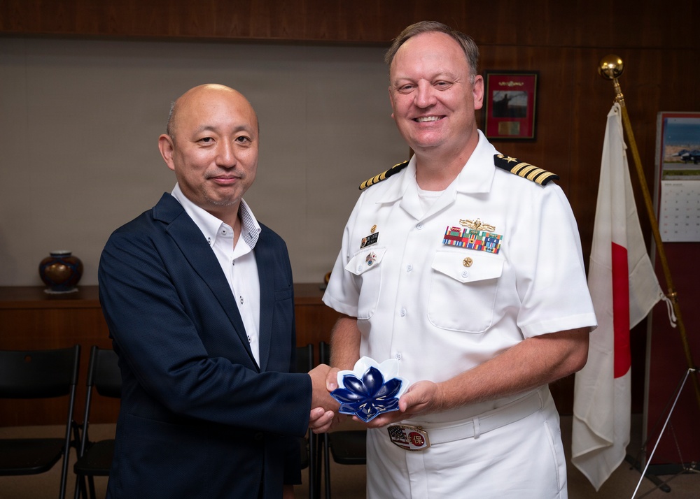 Two men are posing together while jointly holding a blue decorative flower-shaped object, likely during a formal exchange or ceremony. One man is wearing a dark civilian suit while the other is dressed in a white U.S. Navy officer's uniform with gold rank insignia and multiple service ribbons. A Japanese flag is visible in the background, suggesting this may be a bilateral or diplomatic meeting.