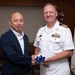 Two men are posing together while jointly holding a blue decorative flower-shaped object, likely during a formal exchange or ceremony. One man is wearing a dark civilian suit while the other is dressed in a white U.S. Navy officer's uniform with gold rank insignia and multiple service ribbons. A Japanese flag is visible in the background, suggesting this may be a bilateral or diplomatic meeting.