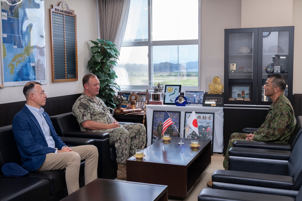 Three individuals are seated in what appears to be a military office, engaged in a formal meeting or discussion. Two individuals wear military uniforms — one in U.S. Navy digital camouflage (NWU) and one in Japanese Ground Self-Defense Force (JGSDF) camouflage — while a third person wears civilian attire. The setting includes both American and Japanese flags on the table, suggesting a bilateral military liaison or diplomatic meeting.
