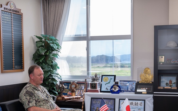 Three individuals are seated in what appears to be a military office, engaged in a formal meeting or discussion. Two individuals wear military uniforms — one in U.S. Navy digital camouflage (NWU) and one in Japanese Ground Self-Defense Force (JGSDF) camouflage — while a third person wears civilian attire. The setting includes both American and Japanese flags on the table, suggesting a bilateral military liaison or diplomatic meeting.