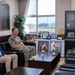 Three individuals are seated in what appears to be a military office, engaged in a formal meeting or discussion. Two individuals wear military uniforms — one in U.S. Navy digital camouflage (NWU) and one in Japanese Ground Self-Defense Force (JGSDF) camouflage — while a third person wears civilian attire. The setting includes both American and Japanese flags on the table, suggesting a bilateral military liaison or diplomatic meeting.