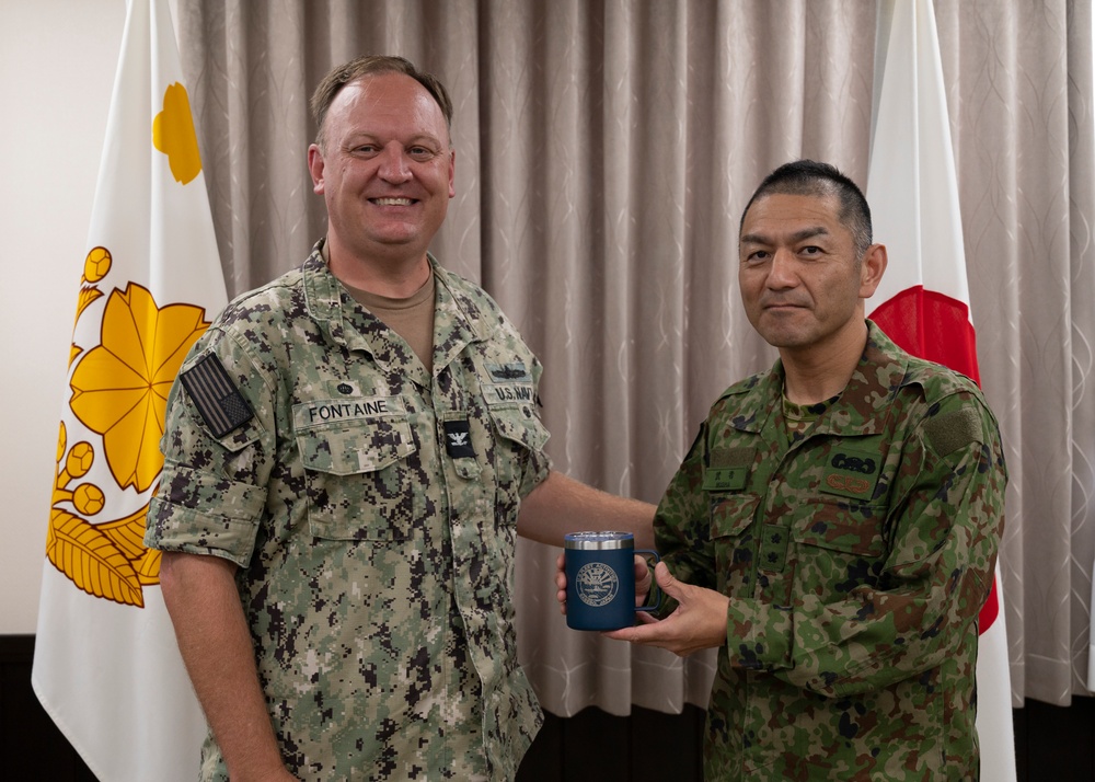 Two military officers, one from the U.S. Navy and one from the Japan Ground Self-Defense Force, are posing together while jointly holding a blue commemorative tumbler/mug. The U.S. Navy officer's name tape reads 'FONTAINE' and he wears a Captain's eagle insignia, while the Japanese officer wears JGSDF woodland camouflage. The flags of both nations' military branches are visible in the background, indicating a formal bilateral military exchange or courtesy visit.