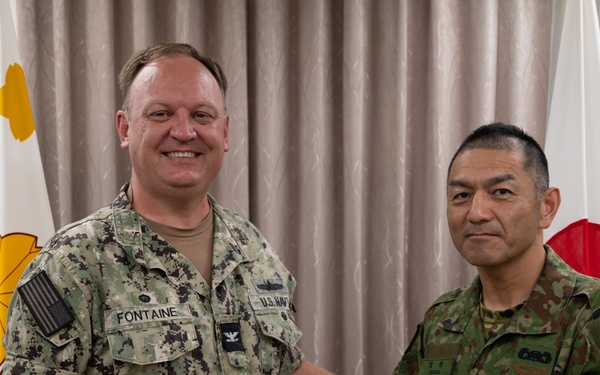Two military officers, one from the U.S. Navy and one from the Japan Ground Self-Defense Force, are posing together while jointly holding a blue commemorative tumbler/mug. The U.S. Navy officer's name tape reads 'FONTAINE' and he wears a Captain's eagle insignia, while the Japanese officer wears JGSDF woodland camouflage. The flags of both nations' military branches are visible in the background, indicating a formal bilateral military exchange or courtesy visit.