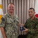 Two military officers, one from the U.S. Navy and one from the Japan Ground Self-Defense Force, are posing together while jointly holding a blue commemorative tumbler/mug. The U.S. Navy officer's name tape reads 'FONTAINE' and he wears a Captain's eagle insignia, while the Japanese officer wears JGSDF woodland camouflage. The flags of both nations' military branches are visible in the background, indicating a formal bilateral military exchange or courtesy visit.