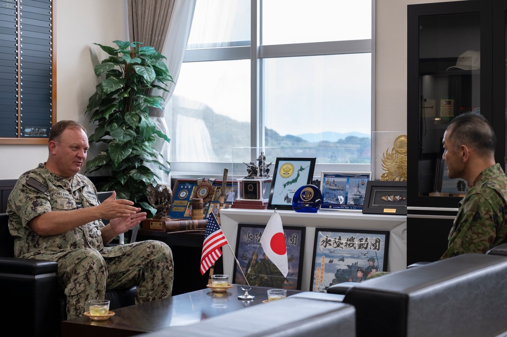 Two military personnel are engaged in a formal meeting or discussion in an office setting. One individual wearing a U.S. Navy camouflage uniform with a name tape reading 'FONTAINE' is speaking, while the other individual in what appears to be a Japanese Ground Self-Defense Force uniform listens. Small U.S. and Japanese flags on the table indicate a bilateral military liaison or coordination meeting.