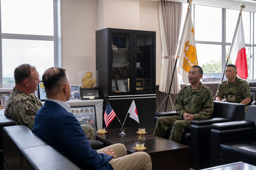 A formal bilateral meeting is taking place between U.S. military/civilian personnel and Japanese Ground Self-Defense Force (JGSDF) officers in what appears to be a commander's office. Small U.S. and Japanese desk flags are prominently displayed on the coffee table between the parties, along with glasses of tea. Full-size Japanese national and unit flags are visible in the background, indicating an official command-level visit.