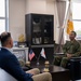 A formal bilateral meeting is taking place between U.S. military/civilian personnel and Japanese Ground Self-Defense Force (JGSDF) officers in what appears to be a commander's office. Small U.S. and Japanese desk flags are prominently displayed on the coffee table between the parties, along with glasses of tea. Full-size Japanese national and unit flags are visible in the background, indicating an official command-level visit.