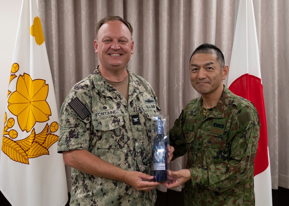 Two military personnel, one U.S. Navy officer and one Japanese Ground Self-Defense Force officer, are posing together while jointly holding a commemorative gift item. The exchange appears to be a formal military courtesy visit or bilateral meeting, evidenced by the flags of both nations visible in the background. The U.S. officer's name tape reads 'FONTAINE' and he wears a Captain (O-6) rank insignia.