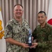Two military personnel, one U.S. Navy officer and one Japanese Ground Self-Defense Force officer, are posing together while jointly holding a commemorative gift item. The exchange appears to be a formal military courtesy visit or bilateral meeting, evidenced by the flags of both nations visible in the background. The U.S. officer's name tape reads 'FONTAINE' and he wears a Captain (O-6) rank insignia.
