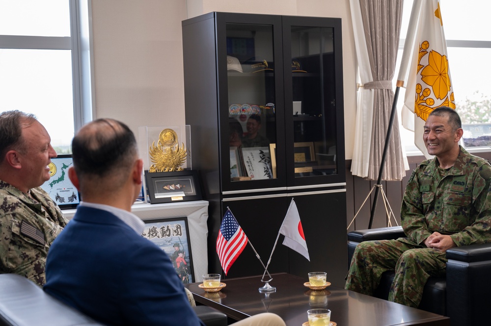 A formal bilateral military meeting between U.S. and Japanese military/civilian personnel, taking place in what appears to be a Japanese military officer's office. Small U.S. and Japanese flags are displayed on the coffee table between the participants, indicating an official diplomatic or liaison meeting. The Japanese officer on the right is smiling, suggesting a cordial and positive exchange.
