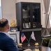 A formal bilateral military meeting between U.S. and Japanese military/civilian personnel, taking place in what appears to be a Japanese military officer's office. Small U.S. and Japanese flags are displayed on the coffee table between the participants, indicating an official diplomatic or liaison meeting. The Japanese officer on the right is smiling, suggesting a cordial and positive exchange.