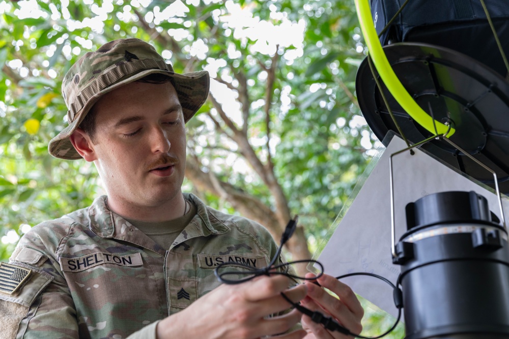 A U.S. Army Sergeant named Shelton is working with what appears to be a tethered drone or aerial system in an outdoor, vegetated environment. He is handling cables or connectors associated with the equipment, which features a yellow-green tether line and black cylindrical components. The setting appears to be a tropical or subtropical location based on the lush green foliage in the background.