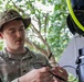 A U.S. Army Sergeant named Shelton is working with what appears to be a tethered drone or aerial system in an outdoor, vegetated environment. He is handling cables or connectors associated with the equipment, which features a yellow-green tether line and black cylindrical components. The setting appears to be a tropical or subtropical location based on the lush green foliage in the background.