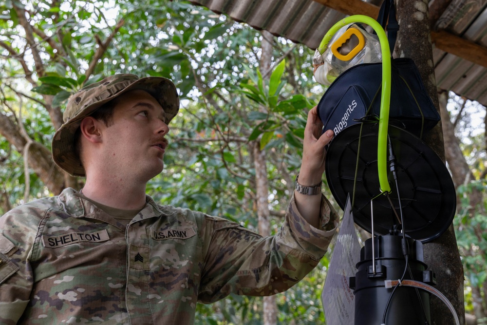 A U.S. Army Sergeant named Shelton is operating or inspecting a piece of equipment mounted on a tree in an outdoor, forested setting. The soldier is wearing MultiCam (OCP) camouflage uniform with a patrol cap and is handling what appears to be a 'Biogents' brand mosquito trap or surveillance device. The setting appears to be a covered outdoor area in a tropical or subtropical environment.