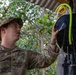 A U.S. Army Sergeant named Shelton is operating or inspecting a piece of equipment mounted on a tree in an outdoor, forested setting. The soldier is wearing MultiCam (OCP) camouflage uniform with a patrol cap and is handling what appears to be a 'Biogents' brand mosquito trap or surveillance device. The setting appears to be a covered outdoor area in a tropical or subtropical environment.