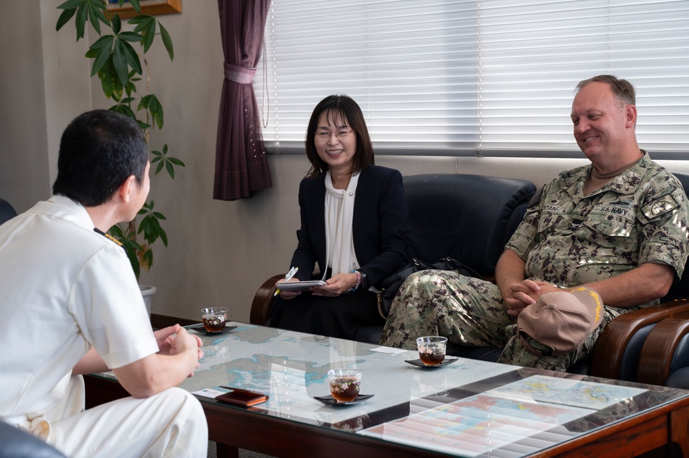 A formal meeting is taking place between a U.S. Navy officer in NWU Type III camouflage uniform and what appears to be a Japanese military or naval officer in white uniform, facilitated by a civilian woman who appears to be acting as an interpreter or liaison. The setting is an office or conference room with a glass-topped table displaying maps underneath. The atmosphere appears cordial and professional, with all parties smiling.