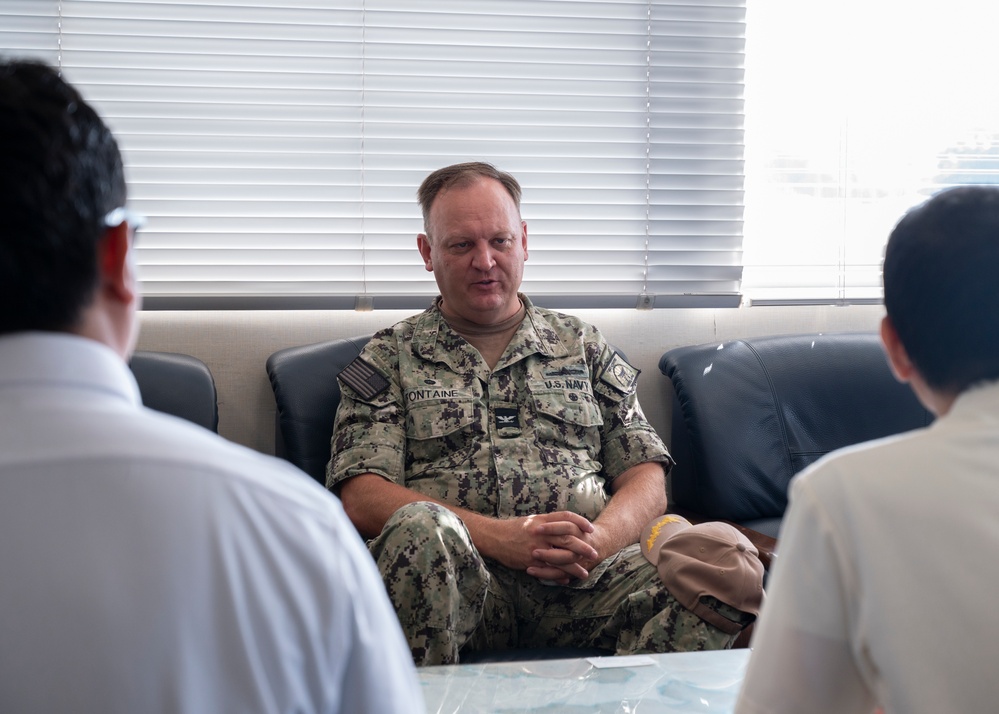 A U.S. Navy officer in NWU Type III camouflage uniform is seated on a leather sofa, engaged in what appears to be a meeting or discussion with two individuals whose backs are visible in the foreground. The officer's name tape reads 'FONTAINE' and the uniform displays 'U.S. NAVY' along with a Captain's eagle insignia and an American flag patch. The setting appears to be an office or conference room with venetian blinds in the background.