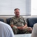 A U.S. Navy officer in NWU Type III camouflage uniform is seated on a leather sofa, engaged in what appears to be a meeting or discussion with two individuals whose backs are visible in the foreground. The officer's name tape reads 'FONTAINE' and the uniform displays 'U.S. NAVY' along with a Captain's eagle insignia and an American flag patch. The setting appears to be an office or conference room with venetian blinds in the background.